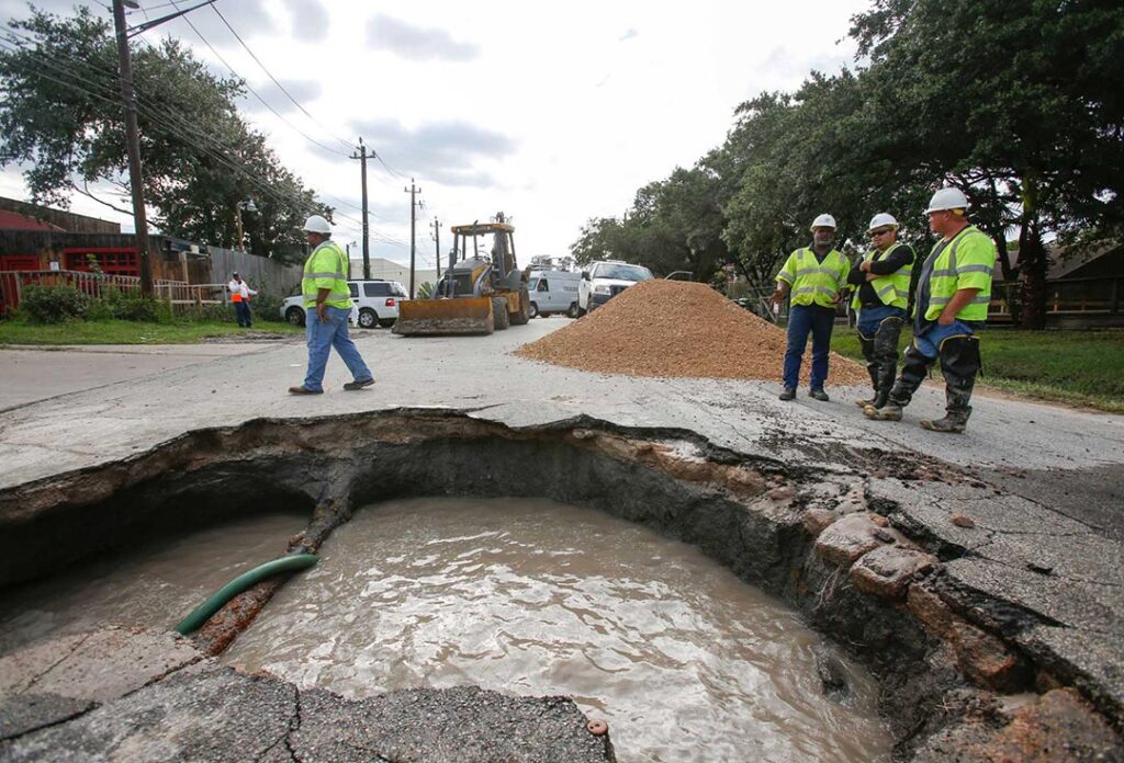 Construction workers stand in front of a hole in a road caused by a water main break