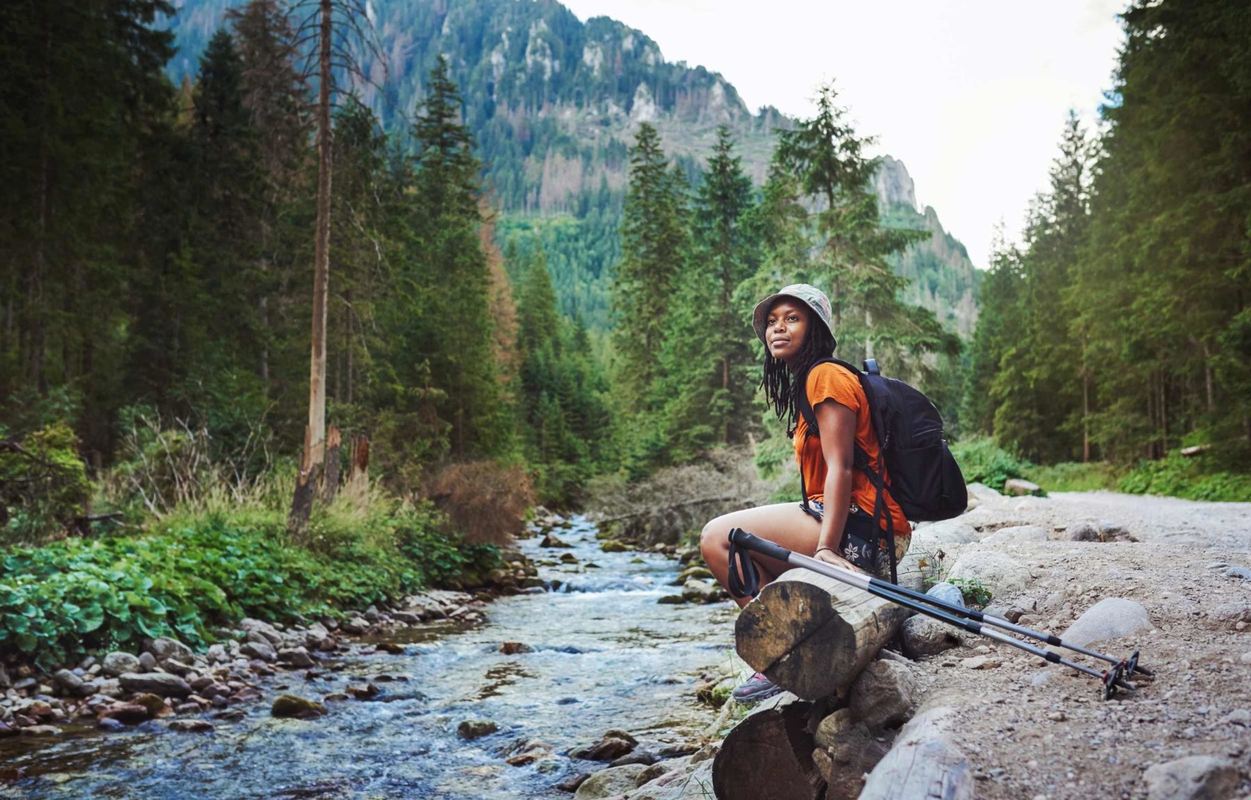 Young woman with hiking gear sits on log next to stream in forest valley